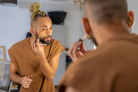 Black male gay applying make up looking mirror standing back to camera. Stylish homosexual man in dressing room doing make-up on face