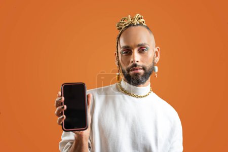 Ein junger lächelnder afrikanisch-amerikanischer schwuler Mann trägt Sweatshirt, das er in der einen Hand hält und zeigt den Smartphone-Bildschirm vor der Kamera isoliert auf orangefarbenem Hintergrund. Lgbtq-Konzept