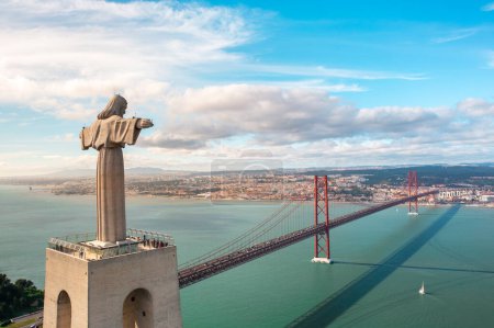 Drone flying around giant sculpture Sanctuary of Christ the King overlooking Portugals capital Lisbon and the 25 de Abril Bridge across Tagus River. Landmarks and infrastructure in Lisbon, Portugal