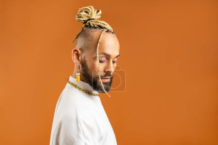 Confident black gay man with bright makeup standing isolated on orange background, dressed white. Exudes sense of pride and individuality. Diversity power of personal style