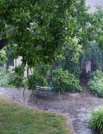 Der Hof im Regen. Es regnet heftig, ein Eichenzweig mit Blättern im Vordergrund, ein verschwommener Blick auf den Bürgersteig und den Weg zum Haus im Hintergrund..
