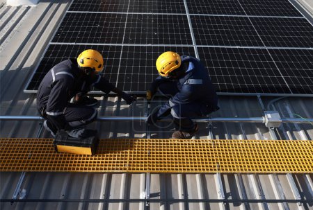 Foto de Dos técnicos con trajes de seguridad PPE y arneses de seguridad caminan sobre el techo solar, el proceso de instalación del panel solar - Imagen libre de derechos