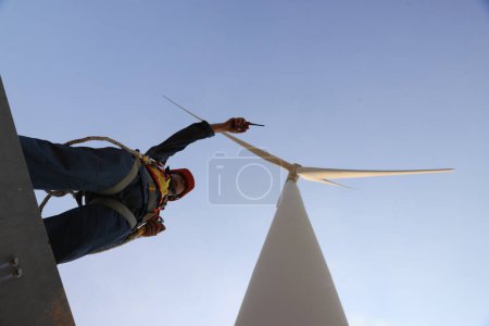 Téléchargez les photos : Specialist wind turbine technician working at the base of the turbine. Wind turbine service technician wearing safety uniform and safety harness working at windmill farm - en image libre de droit