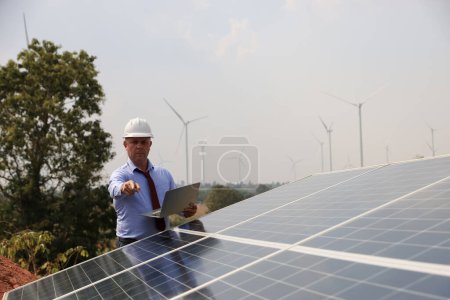 Foto de Un ingeniero masculino sénior inspecciona paneles solares en la granja del molino de viento. Energía limpia. Ingenieros inspeccionan un sistema de paneles solares con una granja de turbinas en el fondo, un concepto de energía renovable - Imagen libre de derechos