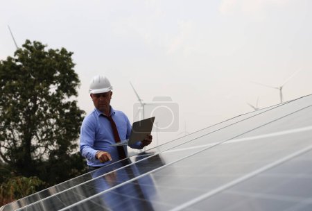 Photo for A senior male engineer inspects solar panels on the windmill farm. Clean energy. Engineers inspect a solar panel system with a turbine farm in the background, a renewable energy concept - Royalty Free Image