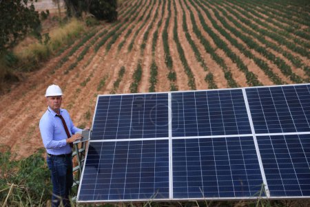Foto de Un ingeniero masculino sénior inspecciona paneles solares en la granja del molino de viento. Energía limpia. Ingenieros inspeccionan un sistema de paneles solares con una granja de turbinas en el fondo, un concepto de energía renovable - Imagen libre de derechos