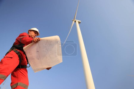 windmill engineer with red safety uniform hold drawing work at front of wind turbine farm 