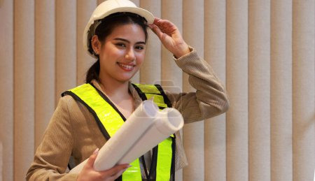  Portrait beautiful female engineer with white safety hardhat and green safety vest holding blueprint drawing