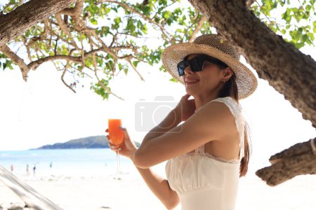 Foto de Una mujer asiática en un traje casual se relaja en la playa junto al mar, sosteniendo un vaso de jugo de naranja y levantando la mano en felicidad. - Imagen libre de derechos