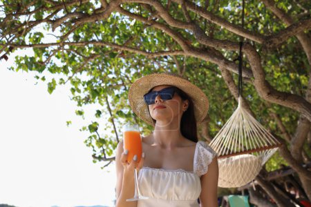 Téléchargez les photos : Une femme asiatique en costume décontracté se détend sur la plage au bord de la mer, tenant un verre de jus d'orange et levant la main dans le bonheur. - en image libre de droit