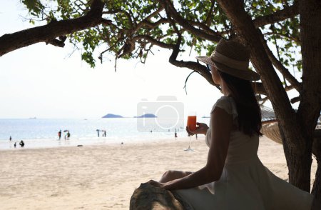 Foto de Una mujer asiática en un traje casual se relaja en la playa junto al mar, sosteniendo un vaso de jugo de naranja y levantando la mano en felicidad. - Imagen libre de derechos