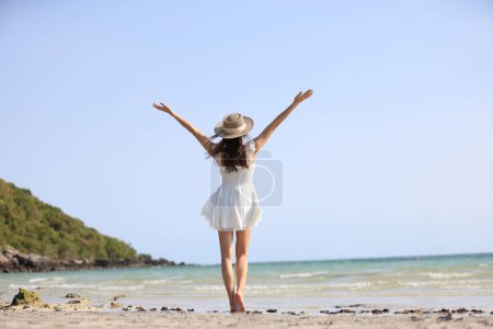Photo for Young attractive ethnic Thai millennial woman posing at suny spreading arms to sides and various postures dressed in white casual clothes stands on the ocean with sandy beach. Love for travel - Royalty Free Image