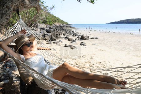 Porträt einer charmanten jungen Asiatin mit Sonnenbrille und glücklichem Lächeln Freizeit auf einer Hängematte Schaukel am Strand Meer und Meer mit blauem Himmel und weißen Wolken für den Urlaub