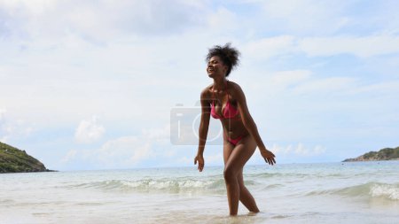 Téléchargez les photos : Une femme noire s'amuse alors qu'elle s'assoit et se détend sur une chaise longue blanche sur une plage tropicale. - en image libre de droit