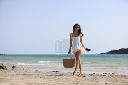 Téléchargez les photos : Joyeux belle femme avec robe décontractée blanche marcher avec pieds nus pour se détendre sur la plage tropicale, vacances d'été et Voyage S'éloigner de tout cela et remplir l'énergie avant de venir au travail - en image libre de droit