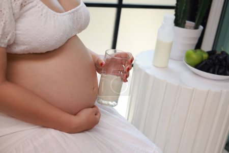 mujer embarazada bebiendo un vaso de agua y comiendo vitaminas en casa. Salud prenatal e hidratación