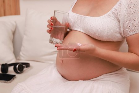 Foto de Mujer embarazada bebiendo un vaso de agua y comiendo vitaminas en casa - Imagen libre de derechos