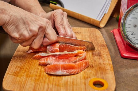Photo for Closeup of hands using sharp knife to slice fresh piece of salmon on wooden cutting board in a well lit and modern kitchen environment. - Royalty Free Image