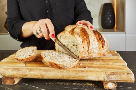 In einer modernen, gut beleuchteten Marmorküche schneidet eine Wirtin vorsichtig einen Laib Brot auf einem Holzschneidebrett. Sie hält ein Messer in der Hand, während sie gekonnt eine Mahlzeit zubereitet.