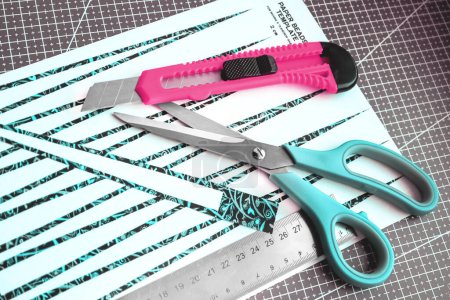 Handmade paper bead templates of green colored floral patterns lying on a cutting mat with pink craft knife and a scissors top down view