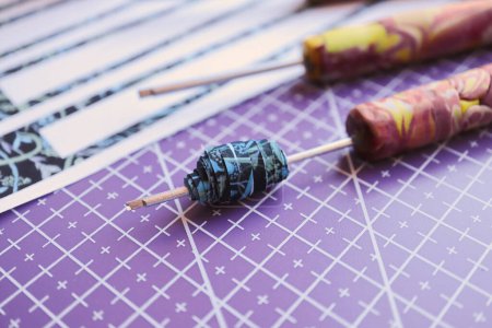Handmade paper bead template of black colored floral pattern being rolled on a rolling tool on a purple cutting mat horizontal side view with bokeh