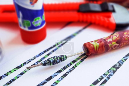 Handmade paper bead template of black colored floral pattern being rolled on a rolling tool with cutting knife and glue stick side view with bokeh