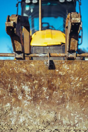 Foto de Primer plano de un tractor amarillo con un gran cubo sucio en un sitio de construcción a la luz del sol brillante. vista frontal - Imagen libre de derechos