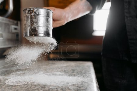 Foto de Close-up of a boy in the kitchen pours and sows flour through a metal sieve into. Home baking concept. The process of making pie, pizza - Imagen libre de derechos