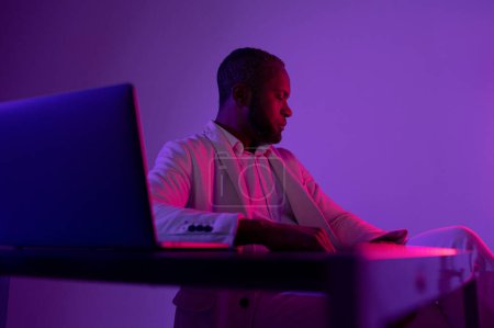 Photo for Black man sitting at the computer in the evening office. IT worker in the evening office. neon light - Royalty Free Image