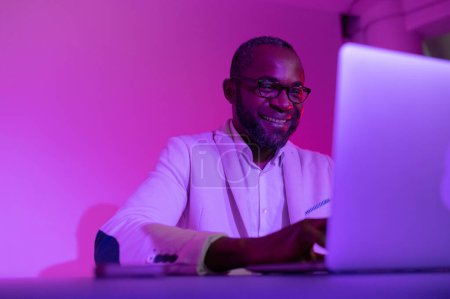 Foto de A black man in a white suit and glasses works at a laptop. a man in glasses works in the office in the night - Imagen libre de derechos