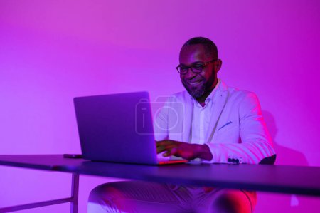 Foto de Un hombre negro con un traje blanco y gafas trabaja en una computadora portátil. un hombre con gafas trabaja en la oficina por la noche - Imagen libre de derechos