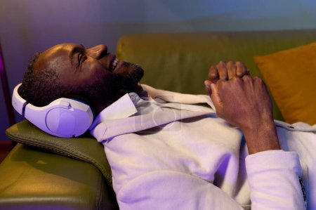 Téléchargez les photos : A black man is lying on the sofa and listening to music. A man in a white suit is lying on a green sofa.music at home - en image libre de droit