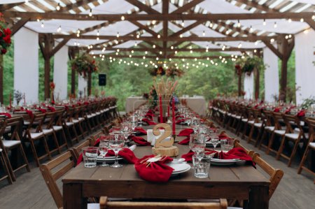 Foto de A large, long, decorated, wooden table and chairs, with dishes, flowers, candles. Pavilion in the forest in nature. Wedding banquet. banquet in nature - Imagen libre de derechos