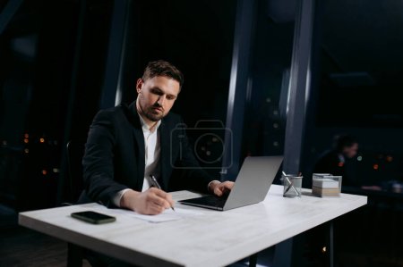 Photo for Young practitioner on the robot to make notes. The male works in the evening in the office - Royalty Free Image