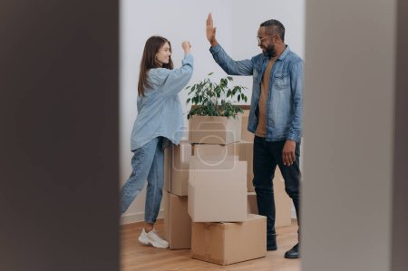 Photo for A young couple high-fives as they stand in the living room, the hallway of their home, next to cardboard boxes of belongings. - Royalty Free Image