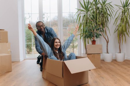 A happy interracial couple, unpacking and laughing on moving day. A happy woman traveling in a cardboard box. African-American man riding a woman in a cardboard box