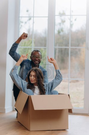 Photo for A happy interracial couple, unpacking and laughing on moving day. A happy woman traveling in a cardboard box. African-American man riding a woman in a cardboard box - Royalty Free Image