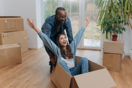 Un couple interracial heureux, déballer et rire le jour du déménagement. Une femme heureuse voyageant dans une boîte en carton. Afro-Américain chevauchant une femme dans une boîte en carton