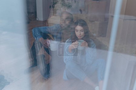 Foto de Una pareja se muda a casa. pareja interracial sentada cerca de cajas de cartón - Imagen libre de derechos