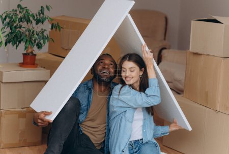 Téléchargez les photos : Un couple emménage dans leur propre maison, ensemble ils font un geste du toit de la maison, assis sur le sol parmi des boîtes en carton - en image libre de droit