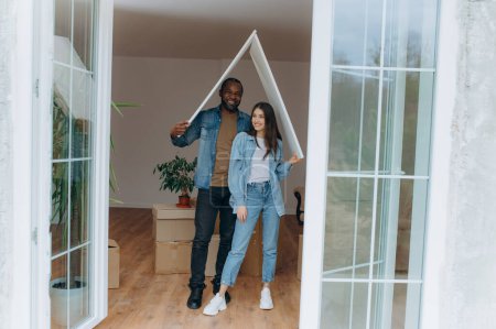 Photo for A couple moves into their own house, together they make a gesture of the roof of the house, sit on the floor among cardboard boxes - Royalty Free Image