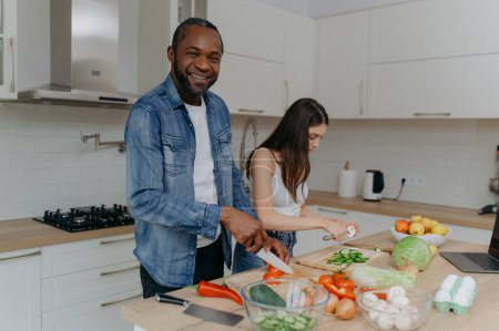 Téléchargez les photos : Un couple interracial heureux qui prépare à manger dans la cuisine. l'homme afro-américain regarde, souriant. - en image libre de droit