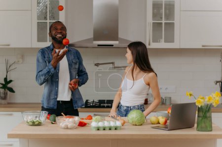 Téléchargez les photos : Une jeune femme heureuse prépare une salade dans la cuisine, un bel homme jongle avec les tomates à proximité. - en image libre de droit