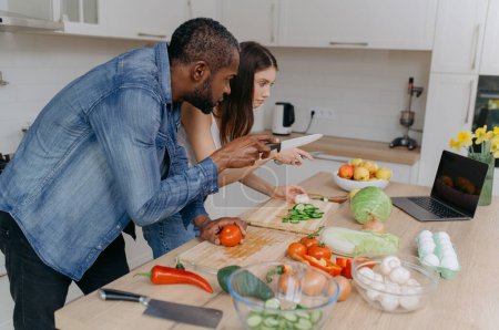 Photo for A married couple cooks together using a laptop, searching for recipes on the Internet. An African-American man and his wife prepare a salad in the kitchen - Royalty Free Image