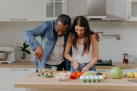 Foto de Una feliz pareja interracial preparando comida en la cocina. Hombre afroamericano y una mujer caucásica preparan una ensalada - Imagen libre de derechos