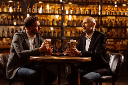 Téléchargez les photos : Amis adultes assis avec des tasses à bière dans un pub. Deux gars joyeux boivent de la bière pression, célèbrent la réunion et sourient. - en image libre de droit