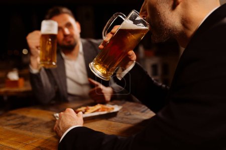 Photo for Adult friends sitting with beer mugs in a pub. Two cheerful guys are drinking draft beer, celebrating meeting and smiling. - Royalty Free Image