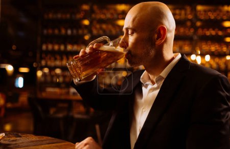 Foto de Un hombre bebe cerveza. Vista lateral del hombre calvo guapo bebiendo cerveza en el pub - Imagen libre de derechos