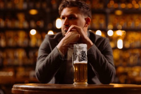 Téléchargez les photos : Triste jeune homme en costume regardant un verre de bière assis à une table dans un pub. Un homme boit après le travail - en image libre de droit