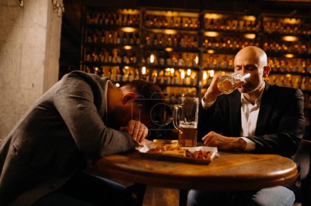 Foto de Un joven con traje duerme cerca de un vaso de whisky y cerveza en una mesa en un pub, otro hombre bebe cerveza. - Imagen libre de derechos